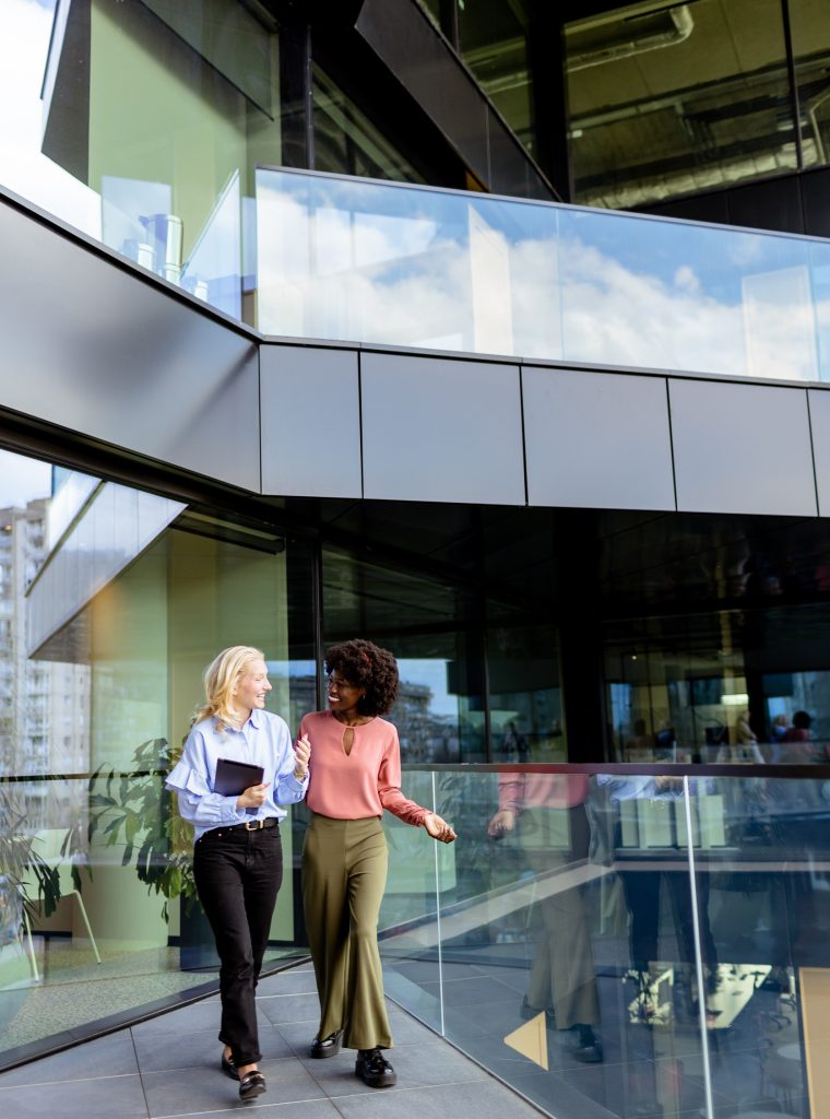 Two colleagues laugh together, one holding a tablet in a sunny, glass-walled workspace.